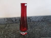 Full view showing tall red glass vase with smooth sides and original paper label near rim.