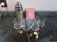 Full view of bar hardware set with shaker, six glasses, cocktail recipe book, bottle opener, corkscrew, and two bottle toppers with pour spouts on dark countertop.