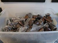 Large clear plastic tote approximately 23" x 15" x 11" filled about two-thirds full with many cone-shaped wood lamp toppers, several visible inside protective plastic bags.