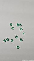 Image shows a scattered collection of 13 small round cut emerald gemstones on a white background, color bright green with high luster.