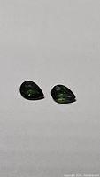 Photo shows two pear-shaped faceted forest green sapphires placed side by side on a plain background.