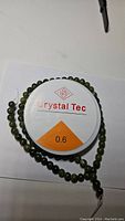 Natural green jade beads on a strand arranged in a circle with a spool of Crystal Tec brand 0.6mm stretchy beading thread in the center on a white background.