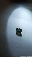 Oval faceted teal blue green sapphire gemstone shown under bright light on white background, highlighting color and cut.