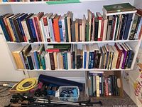 Two shelves filled with various hardcover and paperback books with a box and some exercise items on the floor below.
