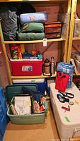 Shelving with three Coleman coolers in red, blue, and white, multiple sleeping bags stacked, and camping supplies on the floor and shelves.