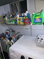 Shelf and utility sink with various cleaning products including Gain detergent, Resolve spray, Clorox wipes, OxiClean, Tide detergent around a large Samsung washing machine.