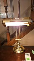 Front and side views of a vintage brass desk lamp showing cylindrical perforated shade and polished brass base set against wooden stair railings.