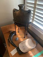 Front and side view of ceramic pilgrim flask on metal stand along with cow horn carvings, wooden bowl and small stone on wood surface.