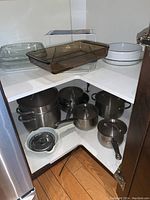 Photo showing multiple glass baking dishes on upper shelves and stainless steel pots with lids and strainer ladle on lower shelf.