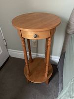 Full view of round wooden side table showing top, single drawer with knob, turned legs, and lower shelf.