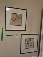 Two framed signed prints hanging on a wall, one smaller above and one larger below. Both frames have decorative metallic borders and beige mats.