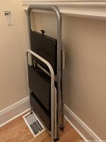 View of two folded metal step ladders stacked against a wall, showing their black plastic steps and silver frame.
