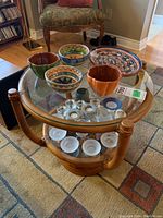 Round wooden table with glass top and lower glass shelf shown with decorative bowls and glass pieces on. Table frame has a natural wood finish and tubular vertical supports.