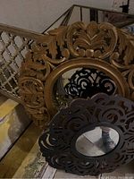 Side view of large gilt wood ornate circular mirror and smaller black cut-out frame mirror stacked against wall