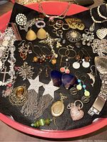 Overview of costume jewelry items arranged on black fabric, showing earrings, rings, pendants, necklace chains, bracelet, and men's watch with metal band.