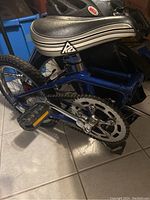 Blue folding bicycle with K2 branded black and white seat visible, bike folded showing pedals and chain.