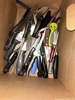 Various kitchen knives with black and colored handles inside a cardboard box, showing numerous utensils mixed together.
