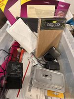 Box containing mixed office supplies with Kodak slide viewer, packs of envelopes, electronic devices, and charger cords.