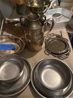 Photo shows various metal dishes including shallow bowls, round plates, silverplate pitcher, and stacked small plates set on a table.