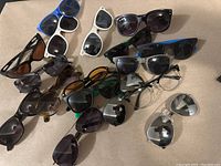 Top-down photo of all sunglasses showing different colors, shapes and sizes scattered on a beige surface