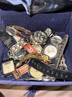 Multiple vintage watches piled inside a dark blue cloth container, showing assorted metal and leather straps and round and rectangular watch faces.