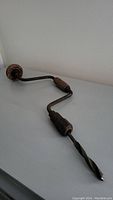 Photo showing the full side view of the antique hand drill with its metal crank, wooden handle, and twisted drill bit.
