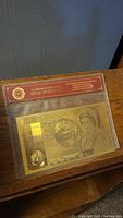 Gold banknote in sealed clear packaging with red Certificate of Authenticity header and embossed design.
