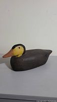 Side view of the wooden decoy duck showing yellow head, black upper neck, orange beak, and brown body with visible texture.