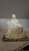 Front and side angled view of the La Pieta resin figurine showing Mary holding Jesus in her lap, mounted on a marble base.