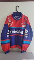 Front view of men's racing jacket in red, blue, teal, and white with large Valvoline logo and multiple embroidered sponsor patches.