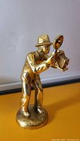 Metal figurine showing front view of man in hat holding antique style camera with bellows and flash attachment, standing on circular base.