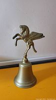 Front angle view of the vintage brass bell featuring the Pegasus winged horse figurine on top against a neutral wall background.