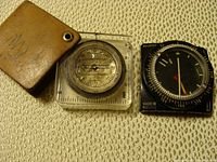 Photo showing a Roots baseplate compass with a brown leather carrying pouch and a black Suunto compass.