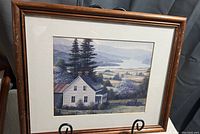 Framed print of a rural house surrounded by trees and fields, with mountains in the distance, presented on an easel.