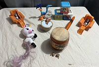 Photo of assorted toys including Looney Tunes characters, unicorn figurine, wooden drum, animal figurines, and wooden stacking blocks on a beige sheet surface.