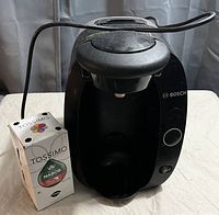 Photo of Bosch Tassimo coffee machine with box of coffee pods on table with curtain background.