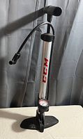 Full view of CCM air pump showing silver body, black handle, attached hose, and pressure gauge at base.