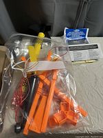 Photo showing the model rocket kit parts in a clear plastic bag, including the main rocket body with black fins, yellow nose cone, and orange launching base components with wires and instructions.