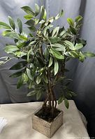 Photo of a faux plant with leafy green branches and brown stems in a beige square textured pot on a flat surface with a gray backdrop.