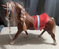 Brown toy horse with red saddle and light blue girth strap, braided mane with blonde and blue strands, white painted hooves.