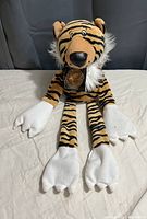 Front view of the tiger plush showing the white fuzzy collar, gold coin necklace, detailed white paws, and embroidered facial features.
