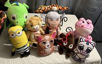 Photo shows a lot of 8 plush toys arranged on a table including cartoon and animal characters.