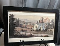 Framed print showing a rural landscape with a house, trees, and hills in muted autumn colors.