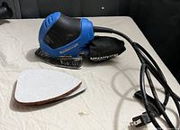 Blue and black Mastercraft electric detail sander with attached dust collection bag and separate triangular sanding pad, displayed on a cloth surface.