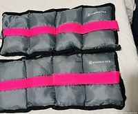 Pair of gray padded ankle weights with pink straps laid flat on a bed.
