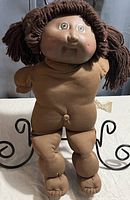 Front view of vintage Cabbage Patch doll showing painted facial features with brown yarn hair and brown fabric body with stuffed legs and arms.