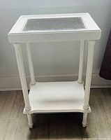 Full view of white wooden side table with glass inset top and lower shelf, standing on tiled floor against white wall.