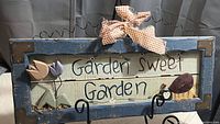 Photo shows wooden garden plaque with blue border and metal corner protectors. There are three wooden tulip flowers and a painted bee figure embellishing the plaque. The phrase 'Garden Sweet Garden' is painted in black on a cream background. A gingham ribbon is tied to the hanging wire loop at top.