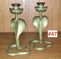 Pair of brass or bronze cobra-shaped candlesticks on a wooden shelf, showing the detailed coiled snakes with raised hoods and smiling faces.