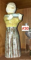 Full view of the ceramic figurative sculpture standing upright on a wooden shelf. Shows figure holding fish with both hands, overall glazed glossy finish with soft earthy colors.